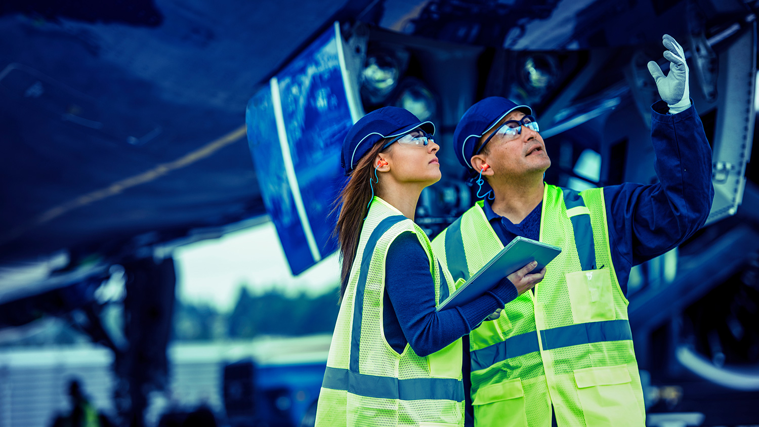 Picture of man and Woman in safety gear looking up, Woman holding a tablet, Man gesturing with left hand.